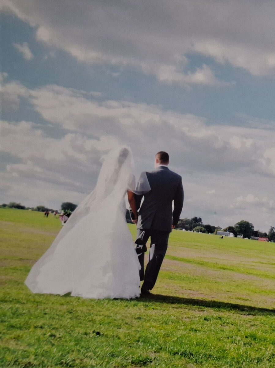 Tania and James walking hand in hand across open parkland, her Sophia Tolli strapless lace gown and long cathedral veil trailing behind.