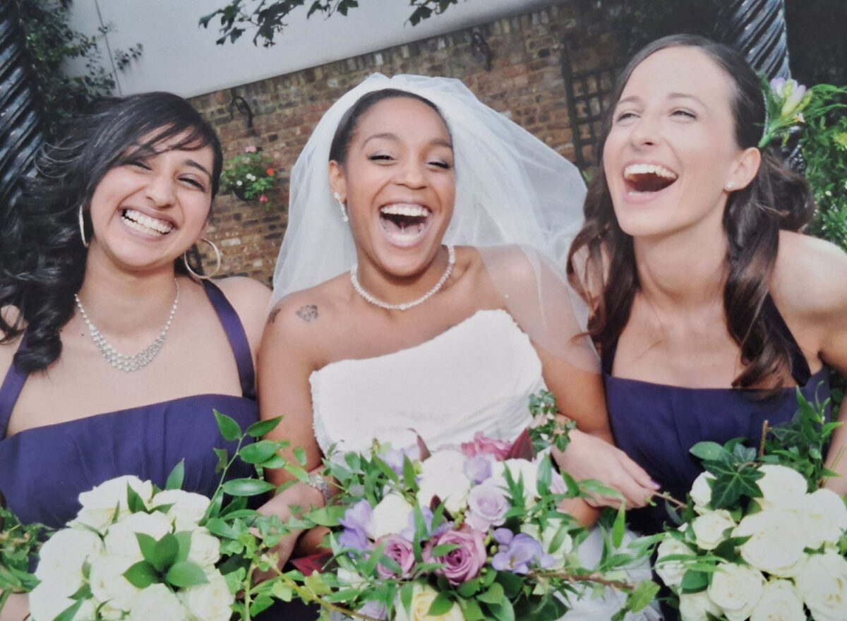 Tania in her strapless Sophia Tolli gown laughing with her two bridesmaids in navy dresses, all holding floral bouquets.