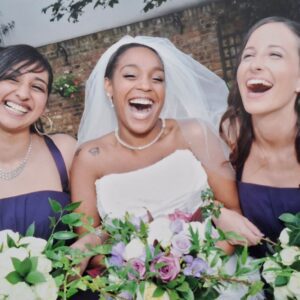 Tania in her strapless Sophia Tolli gown laughing with her two bridesmaids in navy dresses, all holding floral bouquets.