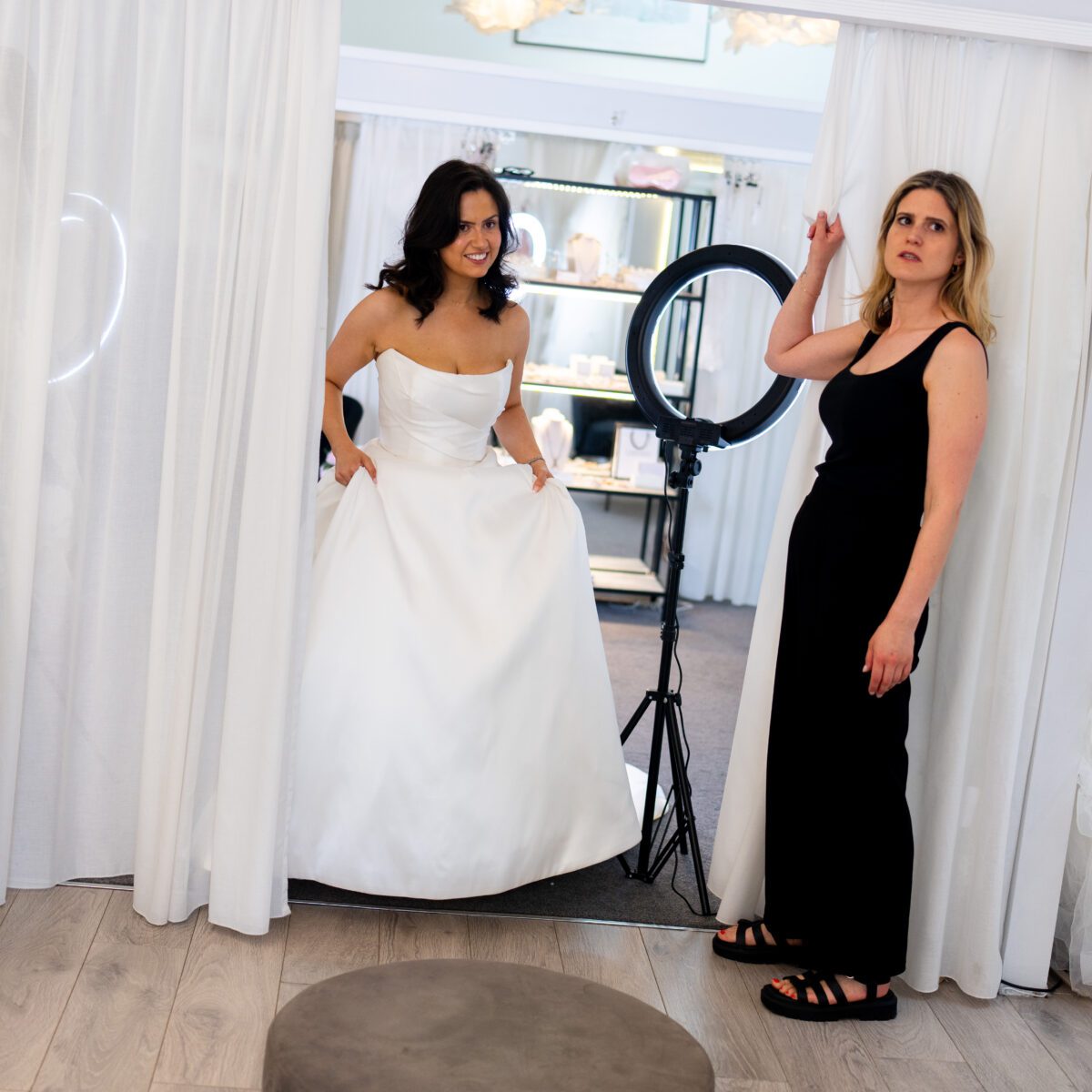 A bride in a satin ballgown steps out of the fitting room curtain while her stylist looks on