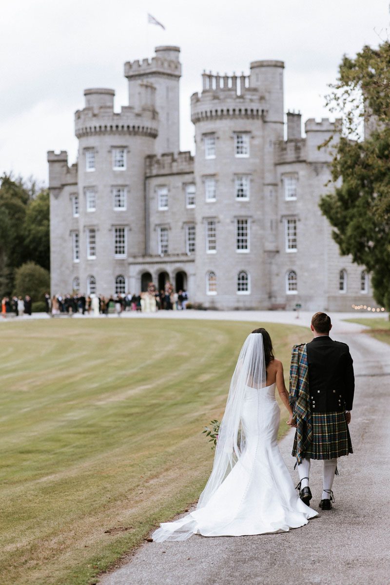 Sophie in her strapless Maggie Sottero mermaid gown and cathedral veil walking hand in hand with Ross in a kilt towards Cluny Castle, Aberdeen.
