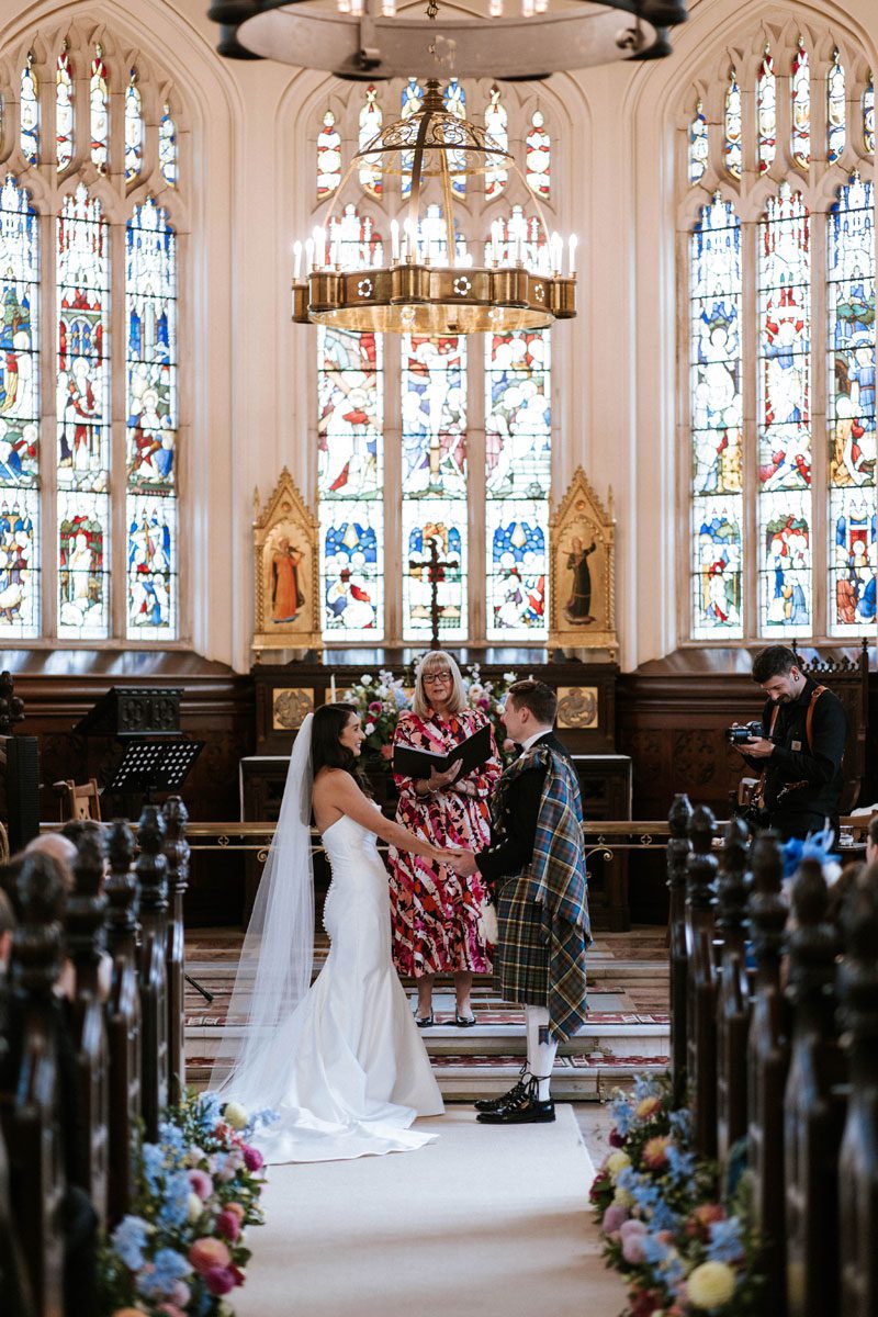 Sophie in her strapless Maggie Sottero mermaid gown holding hands with Ross in a tartan kilt at the altar of Cluny Castle.