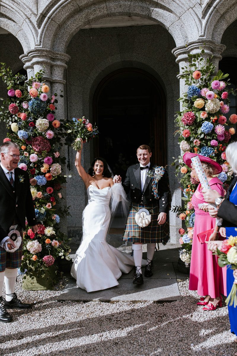 Sophie in her strapless Maggie Sottero mermaid gown raises her bouquet as she and Ross exit Cluny Castle through colourful floral columns.