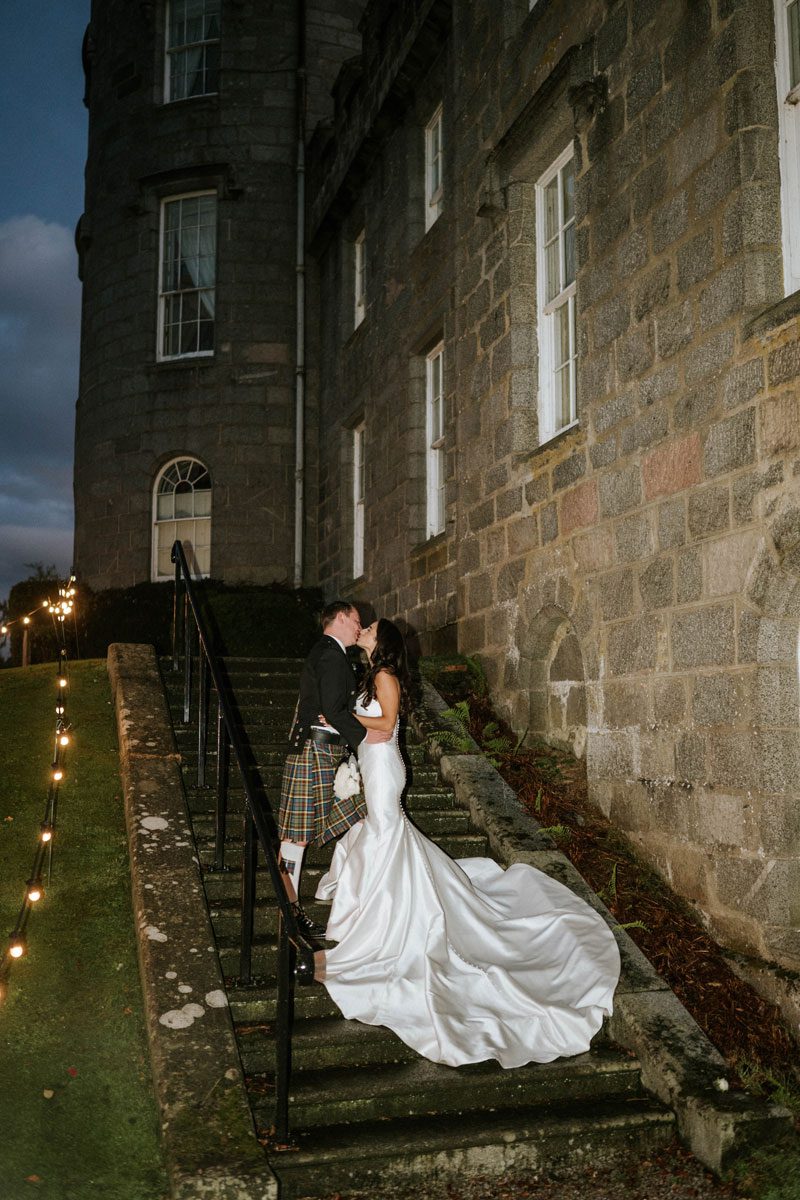 Sophie and Ross share a kiss on the stone steps of Cluny Castle, Sophie wearing a strapless Maggie Sottero mermaid gown with a sweeping train.