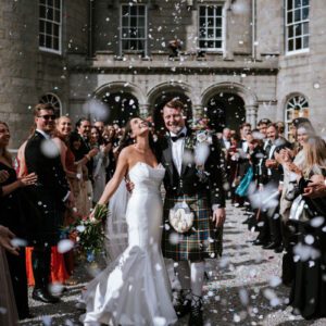 Sophie in her strapless Maggie Sottero mermaid gown laughing in a confetti shower outside Cluny Castle, Aberdeen, with Ross in a tartan kilt.