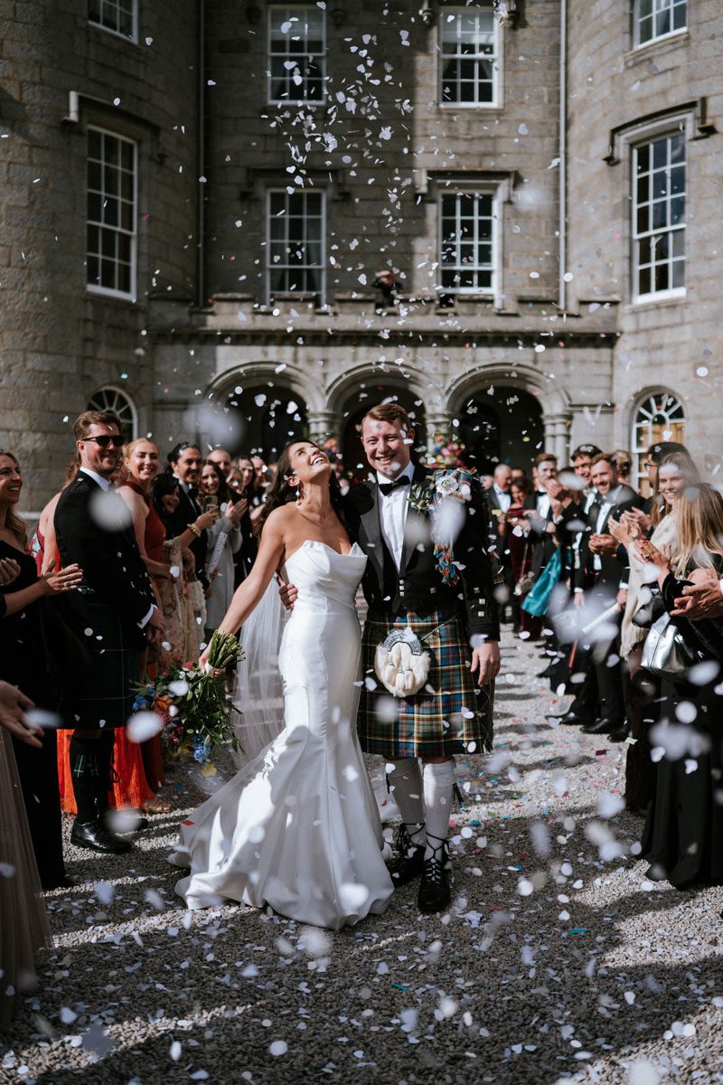 Sophie in her strapless Maggie Sottero mermaid gown laughing in a confetti shower outside Cluny Castle, Aberdeen, with Ross in a tartan kilt.