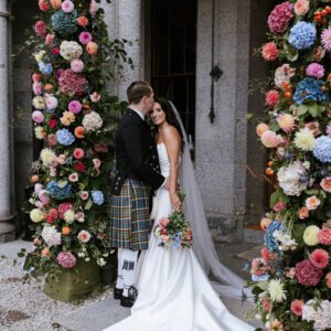 Sophie in her Maggie Sottero strapless mermaid gown with a long veil, held close by Ross in a tartan kilt amid colourful floral columns at Cluny Castle.