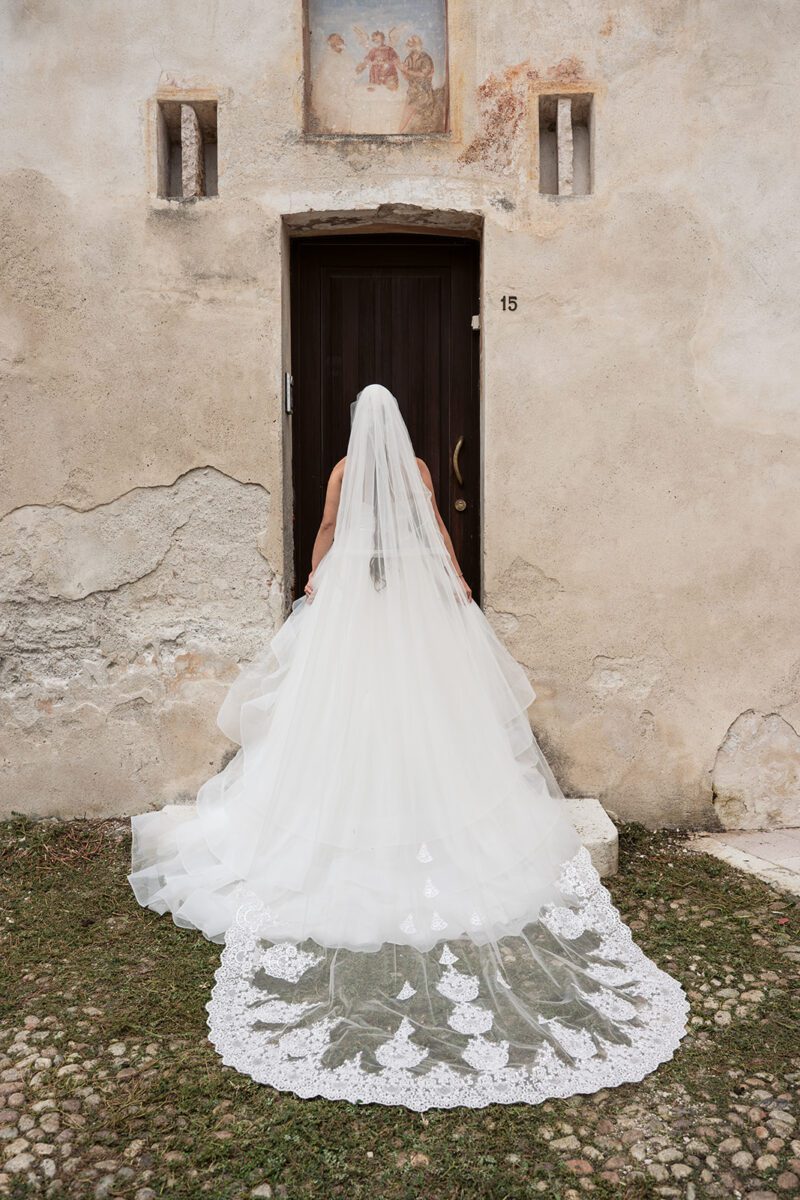The back of Bella's Justin Alexander tiered waterfall gown and lace-trimmed cathedral veil, in front of an old wooden door.