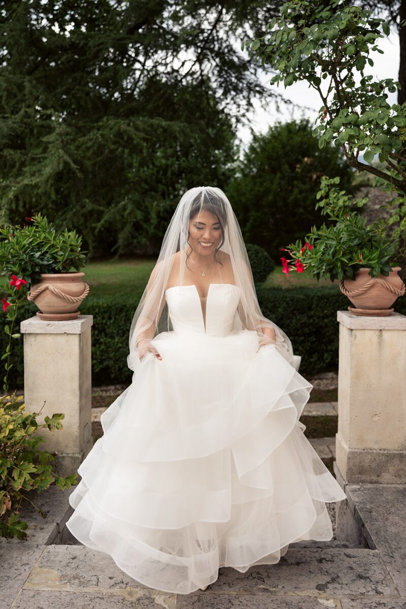 Bella in her Justin Alexander tiered waterfall tulle ballgown and veil, smiling on garden steps in Italy.