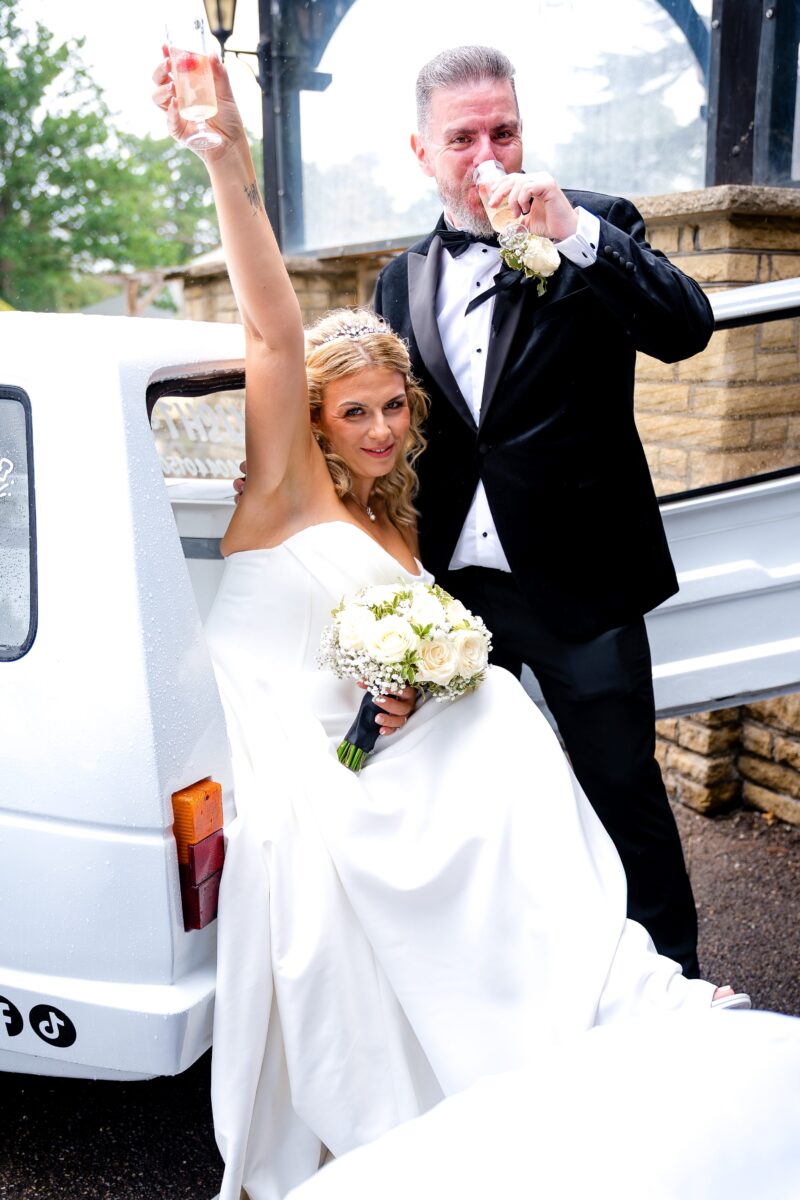 Sarah in her strapless Mori Lee corset gown raising a glass of prosecco beside Anthony, next to the prosecco van at The Manor at Bickley.