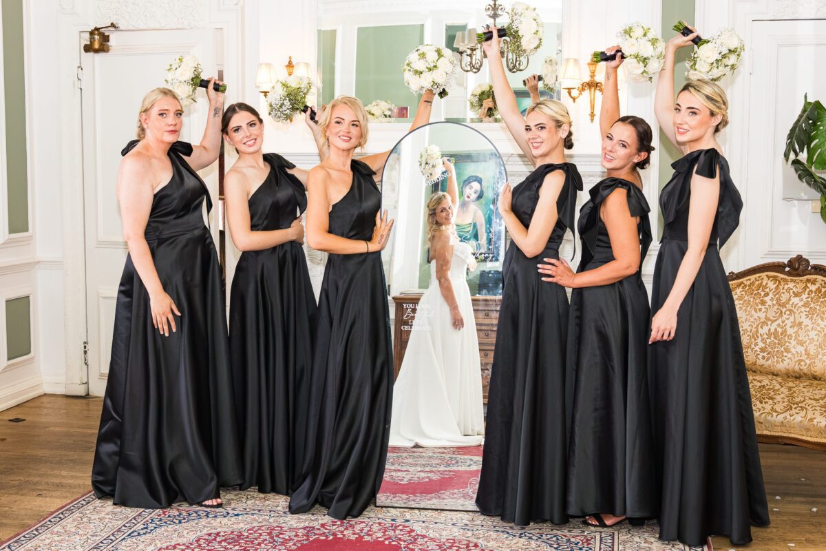 Sarah's six bridesmaids in elegant black satin gowns posing playfully with bouquets raised, with Sarah reflected in a mirror behind them.