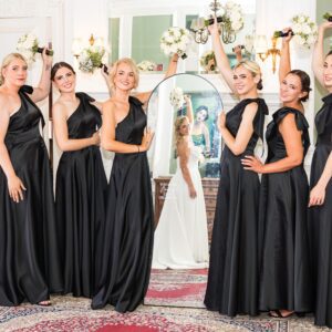 Sarah's six bridesmaids in elegant black satin gowns posing playfully with bouquets raised, with Sarah reflected in a mirror behind them.