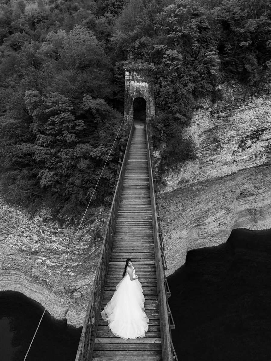 Aerial black and white view of Bella in her tiered waterfall gown on the wooden suspension bridge over Lago del Corlo.