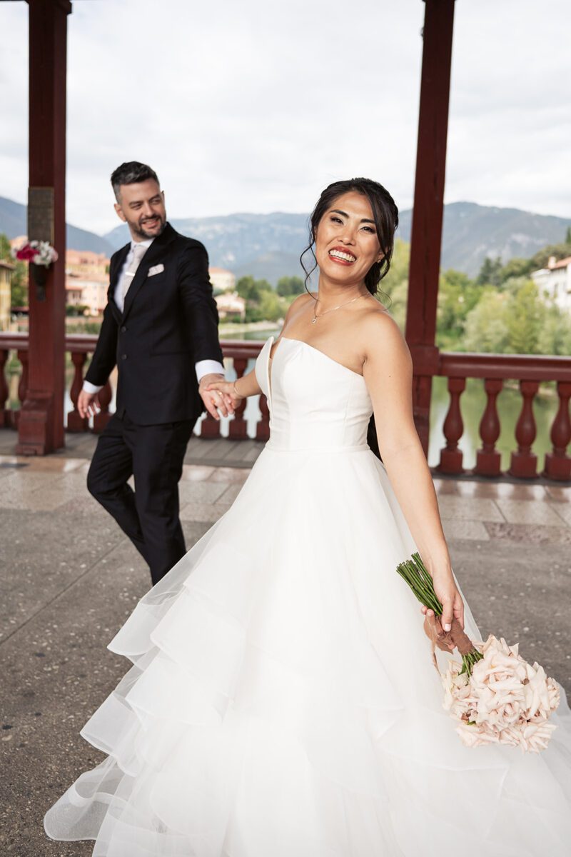Bella smiling over her shoulder in her Justin Alexander tiered waterfall ballgown, holding hands with Alessandro on the Ponte Vecchio, Bassano del Grappa.
