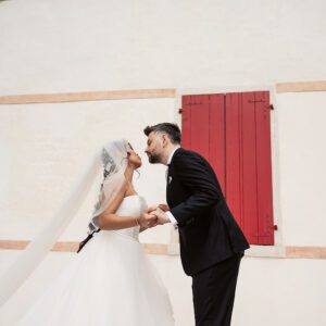 Bella in her Justin Alexander tiered waterfall gown sharing a kiss with Alessandro beside red shutters in Italy.