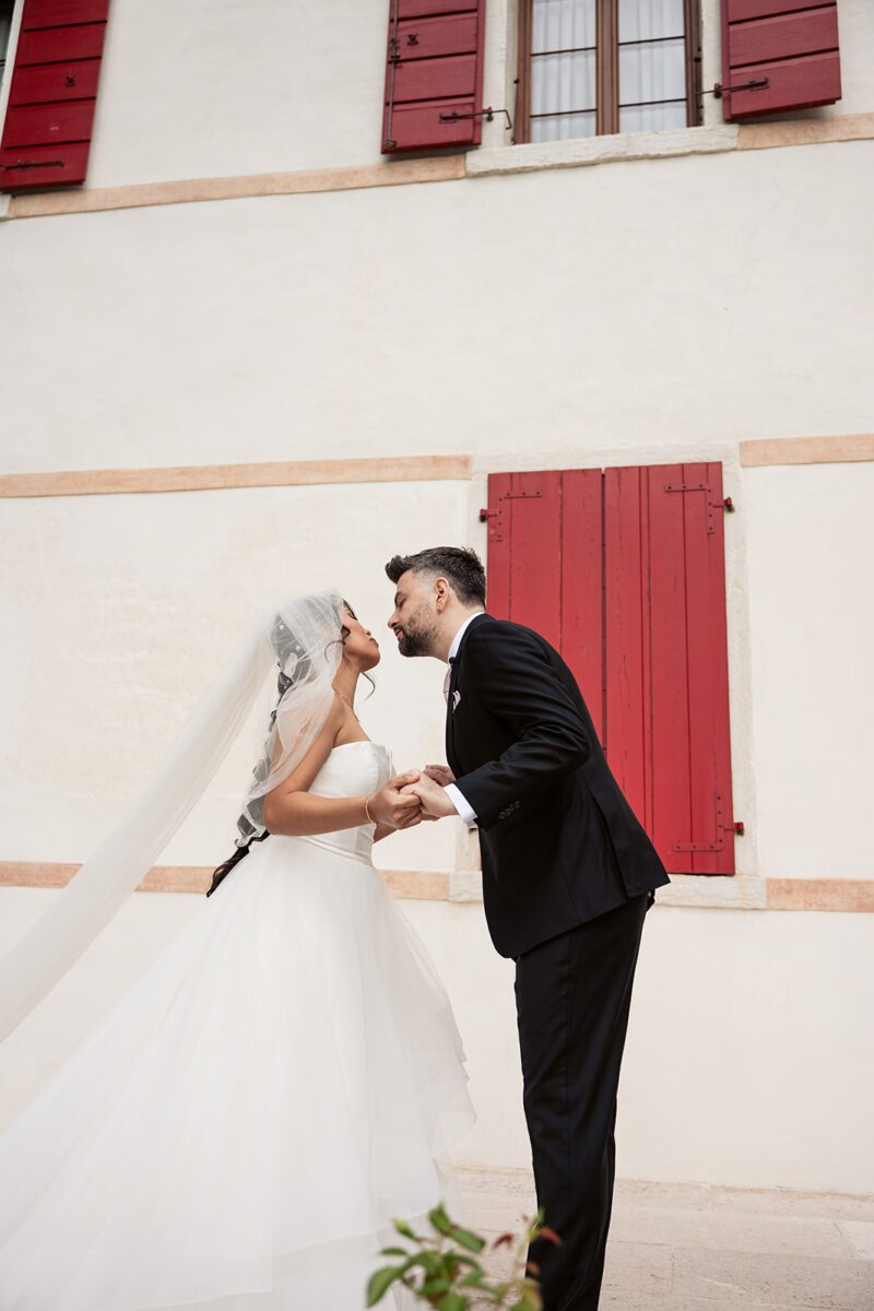 Bella in her Justin Alexander tiered waterfall gown sharing a kiss with Alessandro beside red shutters in Italy.